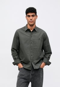 Camisa verde oscura de botones con un bolsillo y mangas largas, hecha de una tela texturizada, combinada con jeans grises.