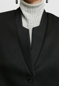 Zwarte blazer met een gestructureerde afwerking en een gelapte kraag, gecombineerd met een lichtgrijze ribgebreide coltrui. Bevat een enkele knoop aan de voorkant.