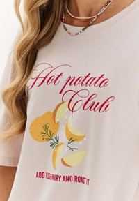 Detailní záběr béžové košile s červeným textem "Hot potato Club" a "Přidejte rozmarýn a upečte to" s ilustracemi brambor a rozmarýnu.