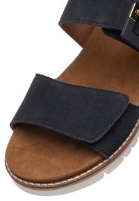 Marineblå ruskindssandal med to brede remme, en polstret tan-farvet indersål og en tekstureret hvid gummisål med syningsdetaljer. Justerbar spænde lukning.