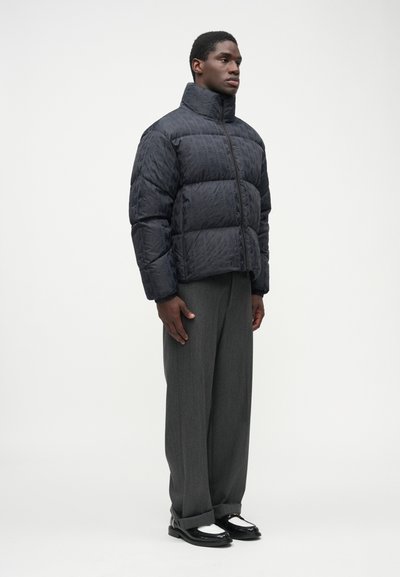 Geacă puffer neagră cu guler înalt și suprafață texturată, asortată cu pantaloni gri și încălțăminte alb-neagră.