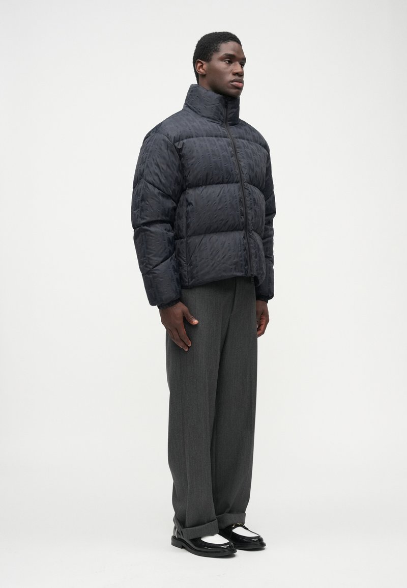 Black puffer jacket with a high collar and textured surface, paired with gray trousers and black and white shoes.