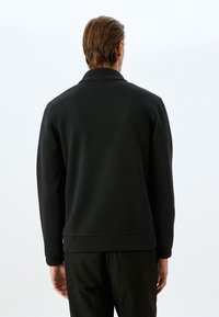 Felpa nera con una texture liscia, polsini e colletto a costine, caratterizzata da un orlo rilassato. Vista dal retro con dettagli di design minimi.