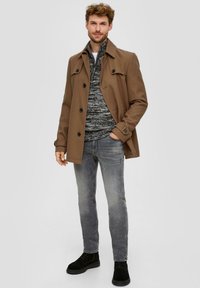 Brauner Trenchcoat über einem grauen Pullover, kombiniert mit grauen Jeans und schwarzen Stiefeln. Die Materialien wirken glatt, mit Knöpfen am Mantel.