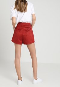 Shorts alti in suede rosso con una texture liscia, tasche laterali e orlo pulito, abbinati a una maglietta bianca e sneakers bianche.