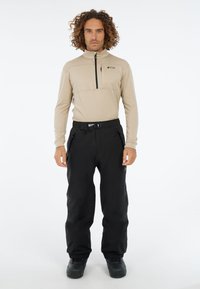 Beige sweater met rits en hoge kraag, gecombineerd met zwarte skibroek. De broek heeft een riem en geribbelde enkels, gedragen met zwarte laarzen.