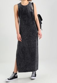 Kobieta w bezrękawnikowej, wyblakłej czarnej sukience maxi, czarnych trampkach za kostkę i niosąca czarny plecak na ramieniu.