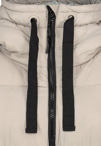 Lichtbeige pufferjack met een zwarte rits, voorzien van twee zwarte koordjes en een quilted textuur. Opvallende hardware omvat logo-drukknoopjes.