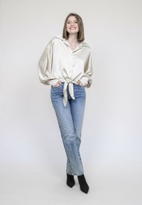 Blouse en satin crème avec un devant à cravate, grand col et manches bouffantes, associée à un jean taille haute bleu clair et des escarpins noirs pointus.