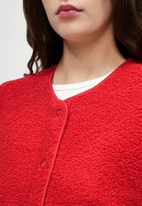 Sarkans vilnas bezpiedurkņu džemperis ar mīkstu tekstūru, noapaļotu kakla izgriezumu un kontrastējošu apdari. Priekšpusē ir pogu aizdare.