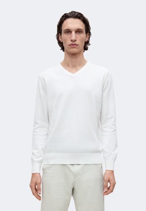 Homme aux cheveux bruns mi-longs portant un pull blanc à col en V et un pantalon clair, debout devant un fond uni gris clair, face à l'avant.