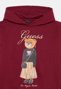 Sweat à capuche bordeaux avec un graphique de ours en dessin animé, portant un pull noir et une jupe plissée dorée, agrémenté d'un sac et de détails de logo.