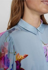 Light blue blouse with a smooth texture, featuring colorful floral patterns on the shoulders. Includes a collared neckline and front zipper.