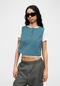 Γυναίκα με μακριά σκούρα μαλλιά, φοράει μαύρα γυαλιά ηλίου, λευκό κοντομάνικο τοπ, τιρκουάζ αμάνικο κοντό γιλέκο και άνετο γκρι παντελόνι.