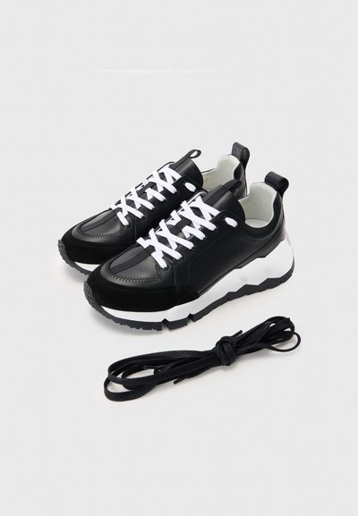 Zapatillas en blanco y negro que presentan una combinación de cuero y gamuza, puntera redondeada, suela gruesa y cordones blancos. Incluye un cordón negro adicional.