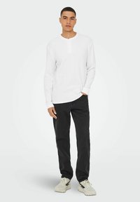 Camisa henley de manga larga blanca con detalles de botones, combinada con pantalones negros y zapatillas beige. La tela parece texturada y casual.