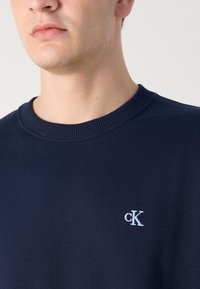 Hajózási kék pulóver bordás kerek nyakkal és egy kis fehér "cK" logóval a mellrészen. Az anyag lágy és textúrált.