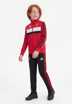 Băiat cu păr roșu zâmbind, purtând un trening roșu și negru Manchester United cu adidași negri, stând pe un fundal alb.