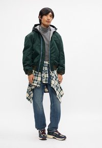 Zelen bomber jakna z sivo kapuco, siva turtleneck majica, karirasta flanelasta srajca zavezana okoli pasu, modri kavbojke in črni tekaški čevlji z belimi detajli.