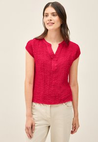 Top corto de manga corta texturizado en rojo con escote de botones. Tejido suave con diseño arrugado, ligeramente ajustado en la cintura. Combinado con pantalones de color claro.