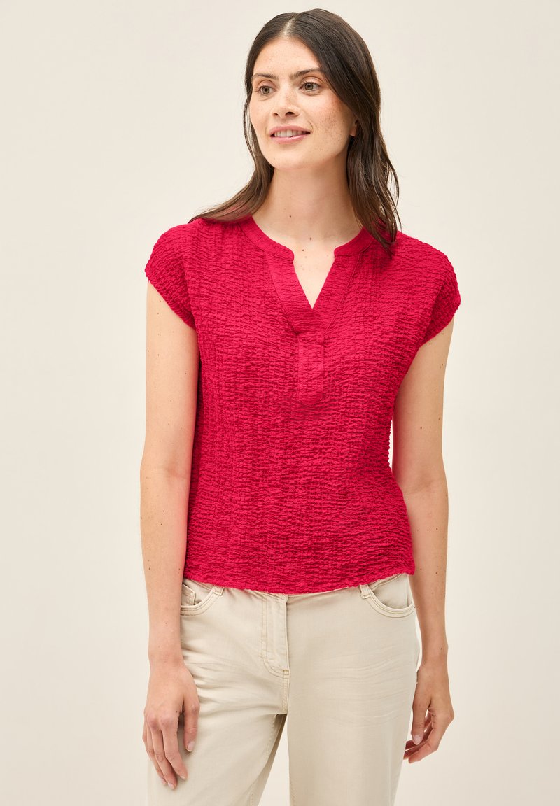 Top corto de manga corta texturizado en rojo con escote de botones. Tejido suave con diseño arrugado, ligeramente ajustado en la cintura. Combinado con pantalones de color claro.