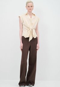Maglietta beige a maniche corte con nodo frontale e colletto, abbinata a pantaloni a gamba larga marroni a vita alta. Il modello si trova su uno sfondo neutro.