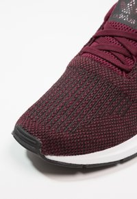 Chaussure de sport bordeaux avec un dessus en tricot texturé et une semelle en caoutchouc noire. Comprend de fines rayures sur le bout et un motif de lacets assorti.