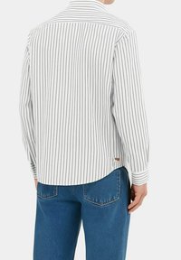 Person iført en hvit langermet skjorte med svarte vertikale striper og blå jeans, vist fra ryggen mot en ensfarget bakgrunn.