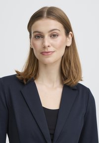 Blazer marine avec un col structuré, une texture douce et une coupe ajustée. Porté par-dessus un haut noir, il présente un revers cranté classique.