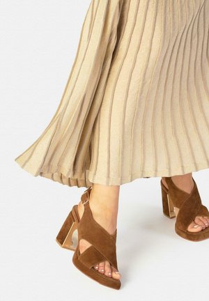 Pies con sandalias de tacón grueso de ante marrón con tiras cruzadas anchas y una falda midi plisada en color beige.