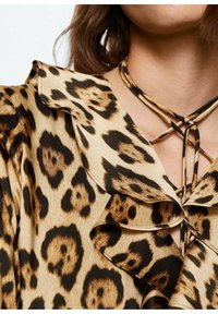 Bluzka w zwierzęcy wzór z leoparda o miękkiej, jedwabistej teksturze, z falbaniastym kołnierzykiem i krzyżującymi się wiązaniami w kolorach brązowym i czarnym.