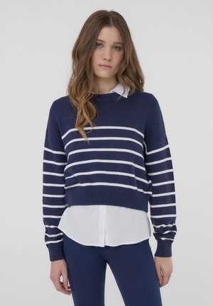 Maglione lavorato a maglia blu navy con righe orizzontali bianche, scollatura rotonda e polsini a coste, indossato sopra una camicia bianca con bottoni.