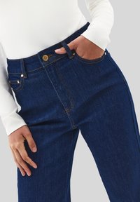 Osoba v tmavě modrých džínech a bílém tričku s dlouhým rukávem, levá ruka v kapse, pravá ruka odpočívá na stehně, neutrální pozadí.