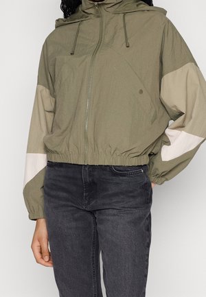 Persoon in een geklede kaki windjack met lichtbeige mouwen en zwarte high-waisted jeans voor een effen achtergrond.