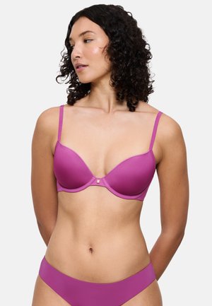 Vaaleanpunainen muotoiltu rintaliivit, joissa säädettävät olkaimet ja kullanvärinen koriste keskellä. Kangas näyttää sileältä ja hieman kiiltävältä, mikä korostaa sen yleisilmettä.