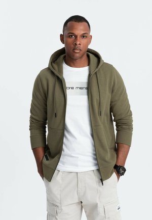 Olijfgroene zip-up hoodie van een textuurstof, met een zak aan de voorkant, gedragen over een witte t-shirt met zwarte tekst. Beige cargobroek.