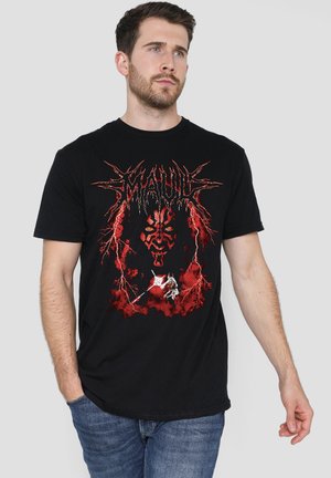 Man with beard wearing black T-shirt with red graphic of a menacing face and figure holding a weapon, standing against a plain background.