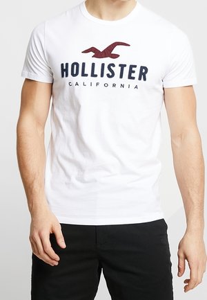 Man draagt een wit Hollister California t-shirt met een rood vogel logo en zwarte broek, staand tegen een effen achtergrond.