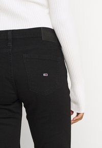 Jeans de mezclilla negros de corte ajustado, con un bolsillo rectangular y una etiqueta texturizada en la cintura. Los detalles incluyen un pequeño logo en el bolsillo.