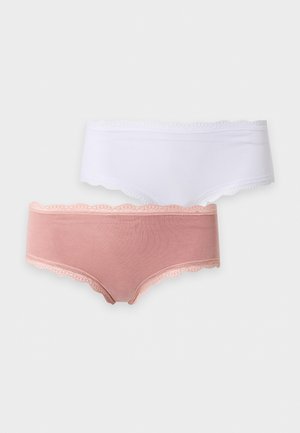 Femme aux longs cheveux foncés, portant un soutien-gorge bleu et une culotte rose bordée de dentelle, debout pieds nus sur un fond blanc uni.
