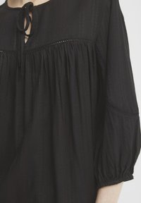 Blusa nera con strisce testurizzate, scollatura a chiave e rifiniture decorative. Presenta maniche voluminose con polsini elasticizzati. Materiale morbido e leggero.