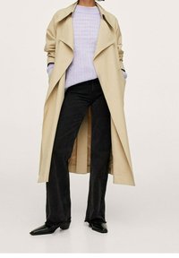 Person wearing a beige long trench coat over a light purple sweater, black flared jeans, and black pointed-toe shoes, standing with hands in pockets.