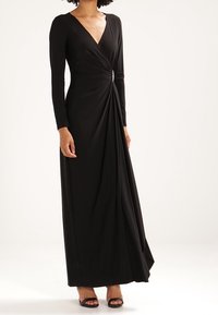 Kvinna klädd i en lång svart omslagsklänning med långa ärmar, V-ringning, sidodetalj med brosch och svarta öppna klackskor mot en enfärgad bakgrund.