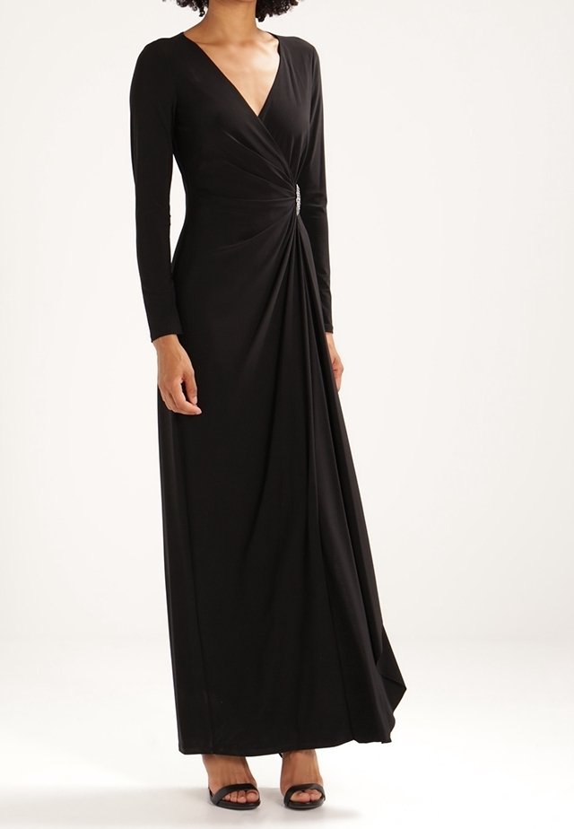 Kvinna klädd i en lång svart omslagsklänning med långa ärmar, V-ringning, sidodetalj med brosch och svarta öppna klackskor mot en enfärgad bakgrund.