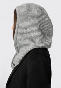 Cappuccio in maglia grigia a costine attaccato a un cappotto nero. Il cappuccio ha una trama morbida che incornicia il viso visto di lato.