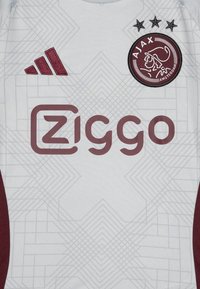 Valkoinen urheilupaita, jossa on viininpunaisia yksityiskohtia, geometrisia kuvioita, brodeerattu logo ja pyöreäreunaisia merkkejä. Adidas-logomerkillä varustettu.