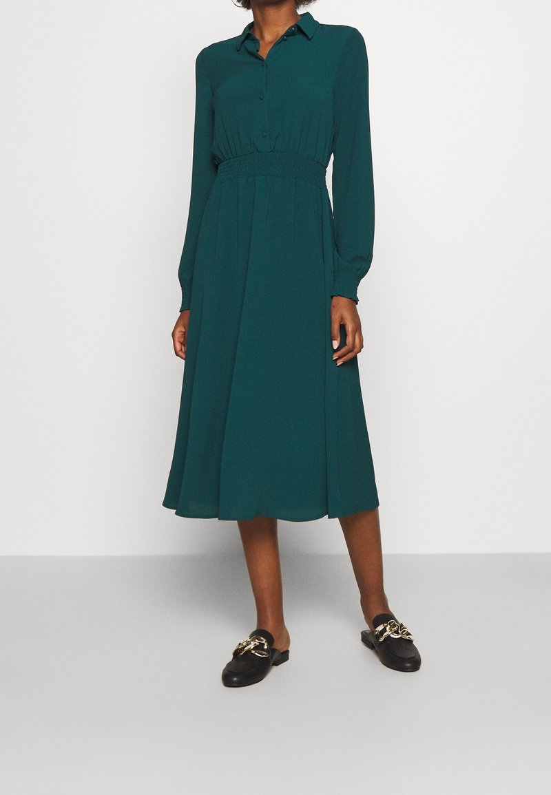 Femme portant une robe midi à manches longues couleur sarcelle avec un col boutonné et des mocassins noirs à enfiler avec un détail chaîne dorée.
