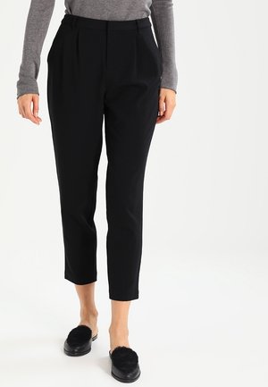 Vrouw draagt zwarte taps toelopende enkellange broek met zijzakken, gecombineerd met zwarte instapschoenen met pluizige bovenkant en een grijs shirt met lange mouwen.