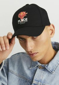Gorra de béisbol negra con un gráfico de una flor y el texto "PLACE INTERNATIONAL ORIGINALS" en la parte frontal, el tejido tiene una textura suave.