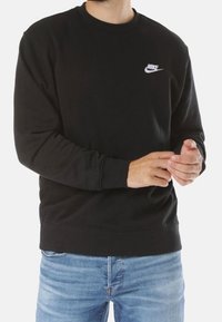 Man som bär en svart Nike-tröja och blå jeans, justerar ärmen med båda händerna.